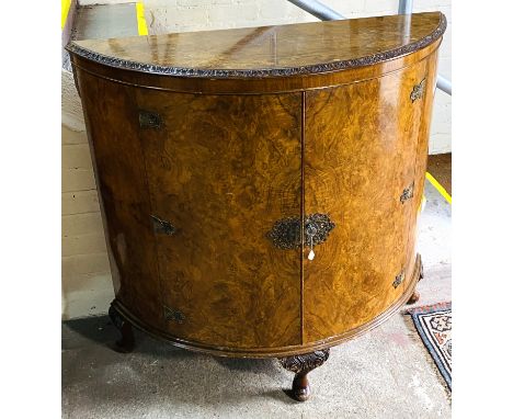 An early 20th century burr walnut demi lune side cabinet with a pair of doors with an internal shelf and brass mounts, raised