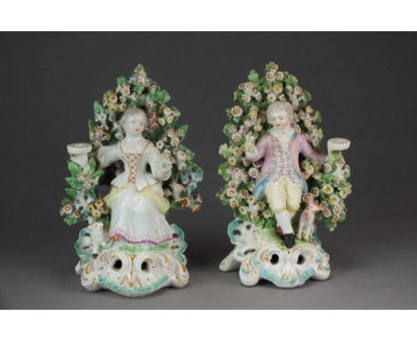 A pair of Chelsea-Derby bocage figural candlestick groups, circa 1765, the female seated beside a lamp, the male beside a hou