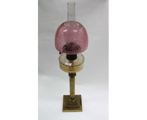 A BRASS CORINTHIAN COLUMN OIL LAMP with a clear glass well and cranberry glass shade 80cm in height 