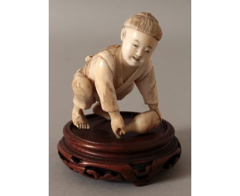 A JAPANESE IVORY OKIMONO OF A BOY, together with a fixed wood stand, the boy bending forward to lift a sake flask from the gr