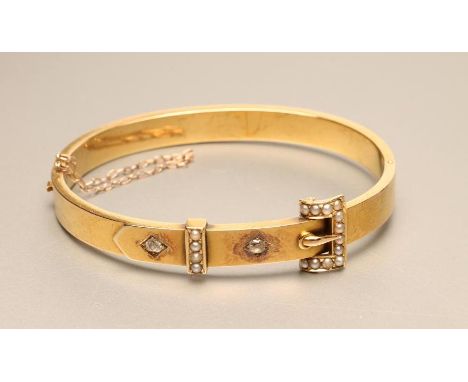 AN EDWARDIAN STIFF HINGED "BUCKLE" BANGLE, the upper section gypsy set with two old cut diamonds and a seed pearl set "buckle