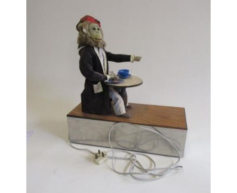 An electrically driven monkey automaton in dressing gown and pyjamas seated at a table, plate, cup, saucer and spoon, eating 
