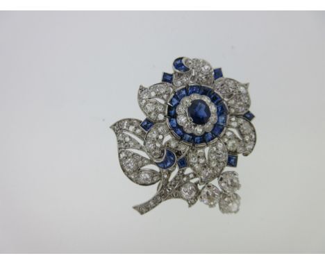 the flower with a central cluster of an oval cut sapphire in a border of ten round old cut diamonds and outer ring of calibré