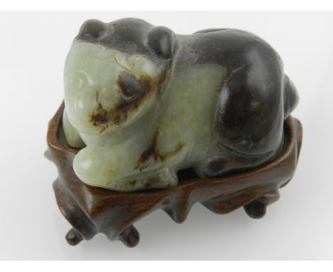 An unusual Chinese mottled green and brown jade carved study of a recumbent ox, raised on a scroll-carved hardwood base. H: 6