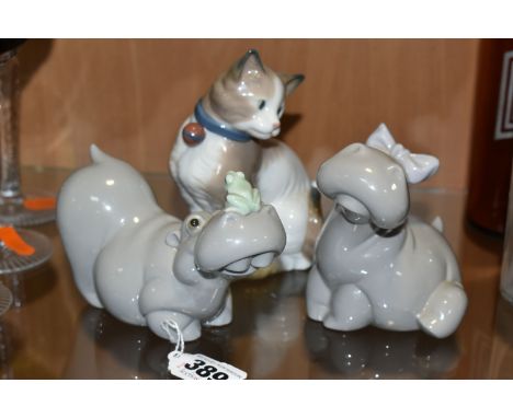 THREE NAO ANIMAL FIGURES, comprising two hippos: one with bow and flower, the other with a frog on its nose and a seated cat,