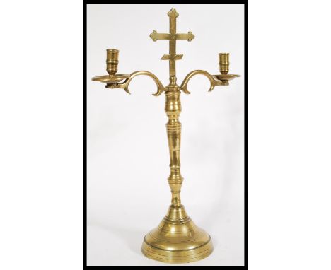 An 18th century ecclesiastical brass candelabra raised on a circular base with twin scroll arms and a religious cross topper.