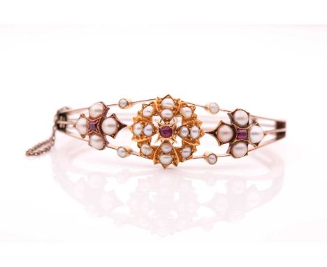 A Victorian 15ct yellow gold, pearl, and ruby bangle, the hinged mount centred with a bright cut gold floral motif centred wi