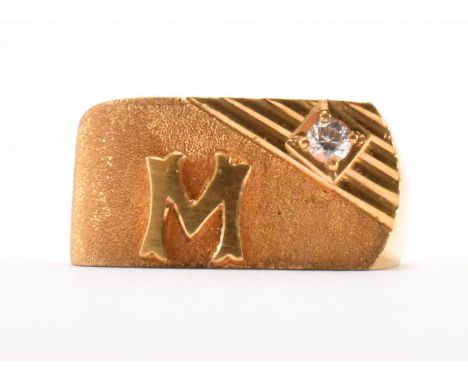 A gold and white stone initial ring. The ring having a square head with raised letter 'M' and set with a round cut white ston