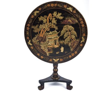 A Chinese black and gold lacquer table, late 19th c, the round tip-up top decorated with two figures before a temple, trees a