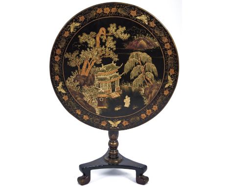 A Chinese black and gold lacquer table, late 19th c, the round tip-up top decorated with two figures before a temple, trees a