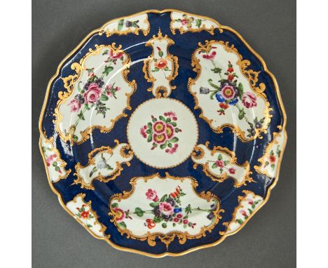 A Worcester scale blue ground plate, c1770, painted with flowers in gilt reserves and lightly upturned shaped gilt rim, 22cm 