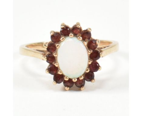 A hallmarked 9ct gold, opal and red stone cluster ring. The ring having a central oval opal cabochon surrounded by a halo of 