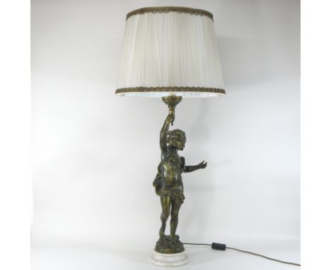 A gilt figural table lamp and shade, in the form of cherub holding a torch aloft, on a white marble base, with a cream shade,