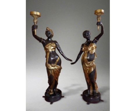 A pair of parcel-gilt bronze figural light stands, each modelled as a semi-clad female, holding aloft a torch in one hand, st