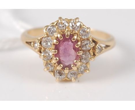 An 18ct gold, ruby and diamond cluster ring, the oval cut ruby within a surround of twelve old cut diamonds, the shoulders ea