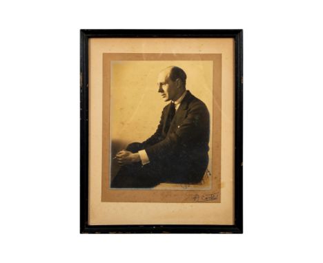 Housed in a black wooden frame with cream color mat. Depicts a gentleman in a dark colored suit, holding a cigarette between 