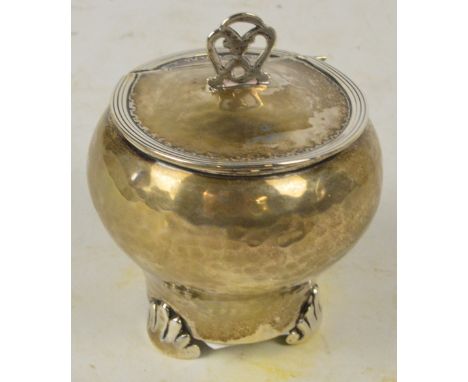 A George V hallmarked silver hammer decorated pot of baluster form with hinged lid and pierced finial, on three stylised supp