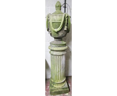 A large weathered composition stone garden urn on fluted column base, the urn dressed with garlands below a flaming finial, e