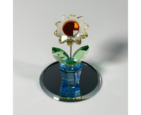 A vibrant, faceted crystal figurine of a yellow sunflower, featuring an orange crystal center, sitting in a blue pot. Swarovs