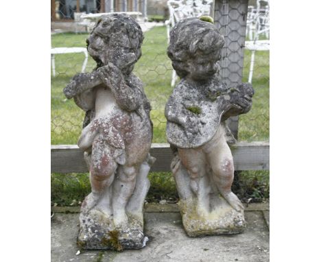 A pair of weathered composite stone figures of putti playing musical instruments, raised on square bases, each approximately 