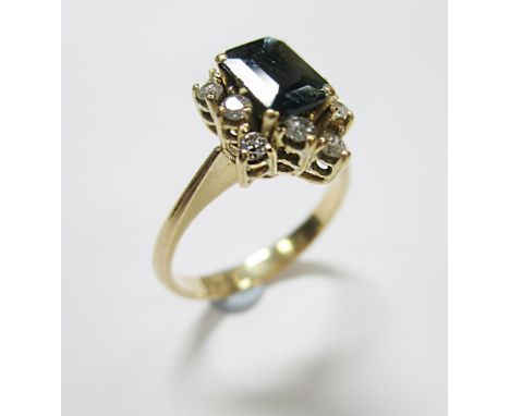 A teal green sapphire and diamond cluster ring, the emerald cut sapphire claw set to an asymmetric undulating border of round