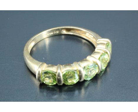 A five stone dress ring, having five 4 mm round green stones, line set between gold batons on plain tapering shoulders, Birmi