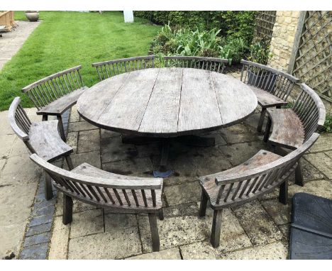 A large circular oak garden table and eight matching curved benches to seat sixteen, the table on four splaying legs, the ben