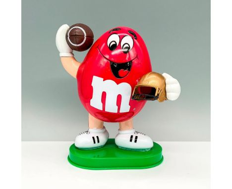 A plastic candy dispenser that depicts a red peanut shaped m and m character holding a football and a gold-colored helmet. Ma