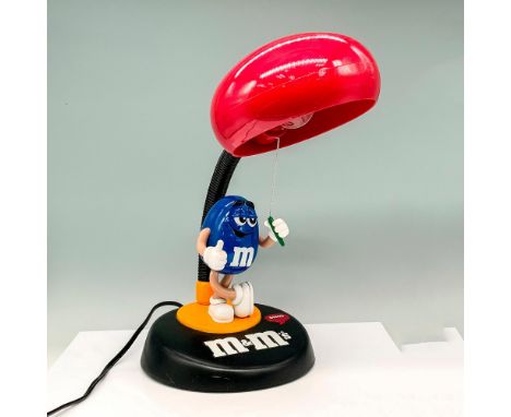 A lamp that has a figure of a blue peanut shaped m and m standing on the base with the lamp shade made to resemble a red m an