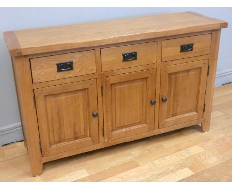 A modern light oak sideboard, comprising of three drawers above three hinged panelled doors, with aged brass effect handles &