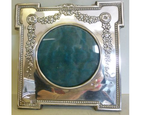 An Edwardian glazed silver photograph frame, having a circular aperture, outset corners, embossed bead and floral garlanded o