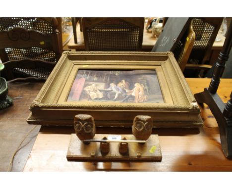 A gilt framed print depicting figures in an interior scene; and a carved wooden desk stand in the form of owls 