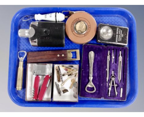 A tray containing vintage tape measure, hip flask, bottle openers, Swiss army knives, Bayer lighter, precision drawing set in