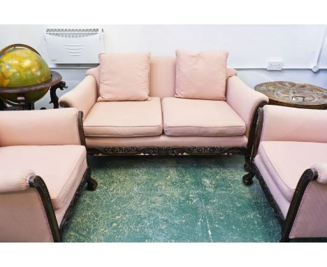 A three piece bergere style suite, upholstered in pink fabric, comprising two-seater sofa and two chairs, carved oak frames w