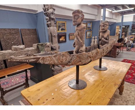 A Timorean dragon canoe, its sides carved with masks detailed in white on a pink ground, two warriors standing inside holding