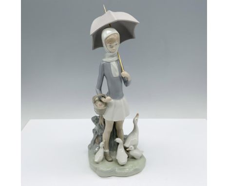 A tall, glossy figurine featuring a girl with a basket in hand and an umbrella, gently observing a family of geese gathered a