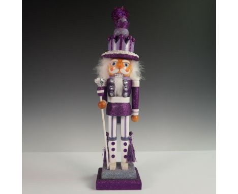 A handcrafted wooden nutcracker figurine adorned in a glittery purple and white ensemble. This regal figure features a high c