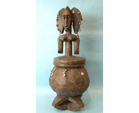 An African carved wood pot and cover, the triple-head finialled lid and bellied pot hung with cowrie shells and supported on 