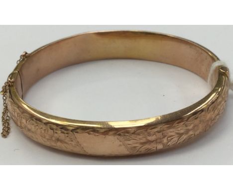 A 9ct gold bangle, approx width 10mm, engraved floral decoration to the top, approx diameter 60mm, hinged with safety chain (