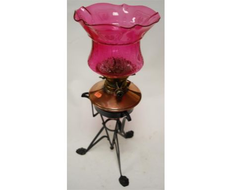An Arts & Crafts oil lamp, having a cranberry glass shade above a copper reservoir, on wrought iron base