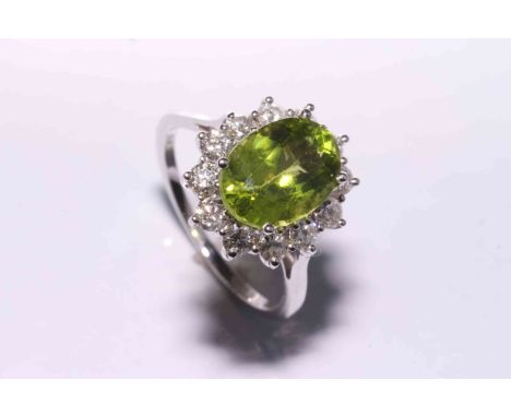 A peridot and diamond ring, the oval cut peridot four claw set, raised within a surround of twelve brilliant cut claw set dia