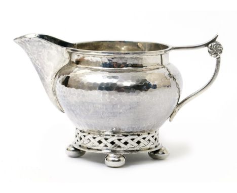 An A.E. Jones silver Celtic cream jug, hammered, shouldered form, on pierced cast trellis band and bun feet, the handle with 