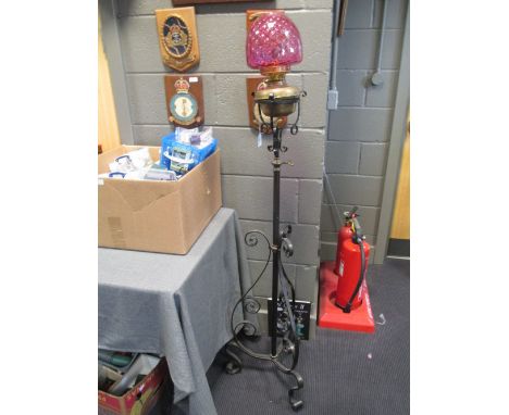 A wrought iron and brass telescopic floor standing frame supporting a brass oil lamp with double wick burner and cranberry gl