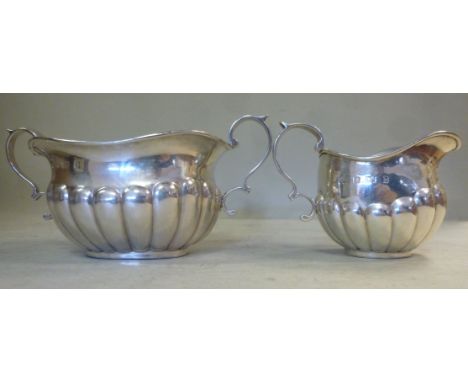 An Edwardian silver cream jug of oval, demi-reeded form with a cast, double C-scrolled handle and a matching twin handled sug