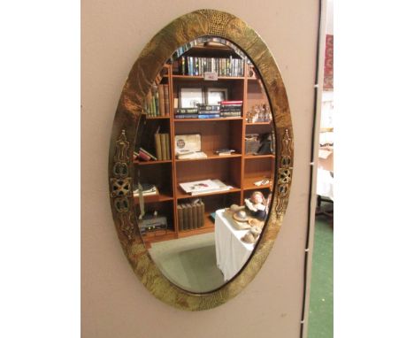 Oval bevelled wall mirror in the Arts and Crafts style with a beaten brass frame and applied brass mounts to either side and 