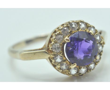 A hallmarked 9ct gold cluster ring set with a round cut central amethyst stone within a halo of round cut white stones. Hallm