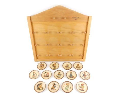 A set of thirteen Goebel Hummel trinket dishes, 8cm diameter, together with a light oak wall hanging display board, 46cm x 45