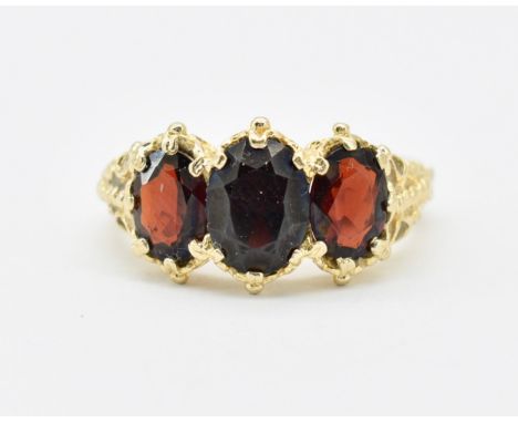 A hallmarked 9ct gold and garnet three stone ring. The ring having three oval cut garnets set within a pierced and scrolled m