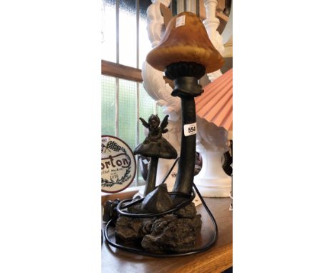A modern cast resin table lamp depicting a fairy relaxing upon a toadstool with mottled amber glass mushroom form shade