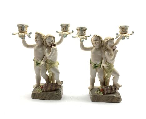  Two 20th century Continental porcelain two branch candelabra modelled as two cherubs playing pan pipes with a cornucopia ove